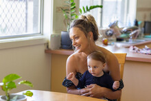 Young Mother Sitting At Kitchen Table With Baby On Lap