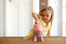 Blonde Girl Wearing Yellow Blouse Putting Coins In The Pink Piggy Bank With Coins On The Table