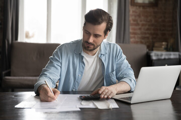 Serious handsome computer user man doing accounting job, checking paper bills, receipts, writing notes in documents, using calculator, counting budget, tax, rent, mortgage fees. Finance management