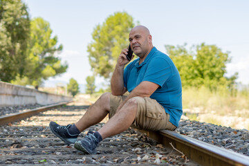 Overweight middle-aged man sitting on the train track talking on the phone.