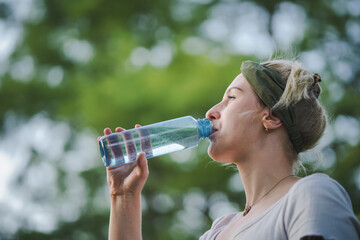 closeup o f a young healthy alternative & spiritual looking woman drinking fresh detox water from a blue plastic bottle outside in the park with a green refreshed nature forest background