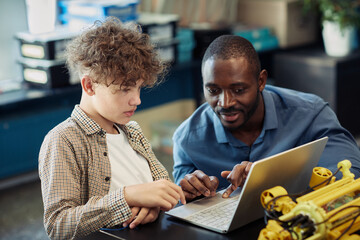 Wall Mural - Portrait of black male teacher helping young boy building robot during engineering class in school and using laptop for programming together