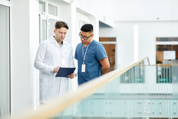 Waist up portrait of two doctors looking at clipboard while standing at balcony in modern clinic interior, copy space