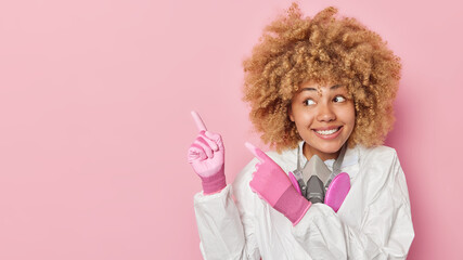 Positive curly haired woman wears white biohazard suit and protective gloves points at upper left corner demonstrates place for your advertisement isolated over pink background. Health care worker