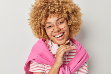 Wall Mural - Beautiful curly haired woman smiles broadly keeps hand under chin focused forward expresses positive emotions wears big spectacles striped t shirt pink pullover tied over shoulders uses headphones