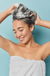 © WHstudio Leushin N - Vertical shot of young Asian woman with healthy skin enjoys showering applies shampoo on wet hair washes head stands in bathroom wrapped in white soft towel. Hair care and daily hygiene concept