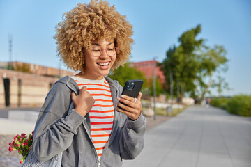Wall Mural - modern millennial girl with curly hair strolls outdoors holds mobile phone chats online makes shoppi