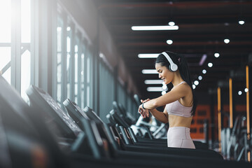 Happy beautiful young asian woman running on treadmill and listening to music via headphone during sports training in a gym.