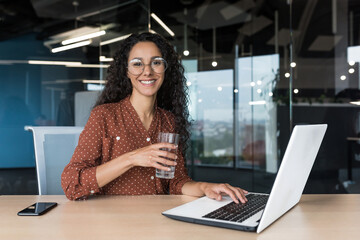 Wall Mural - Portrait of happy and successful business woman inside modern office building, female worker smiling and looking at camera, holding glass of clean water, working on laptop at desk