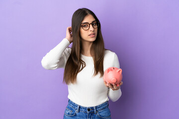 Wall Mural - Young caucasian woman holding a piggybank isolated on purple background having doubts