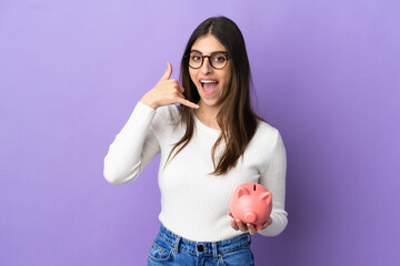 Wall Mural - Young caucasian woman holding a piggybank isolated on purple background making phone gesture. Call me back sign