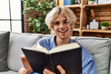 Poster - Young blond man reading book sitting on sofa at home