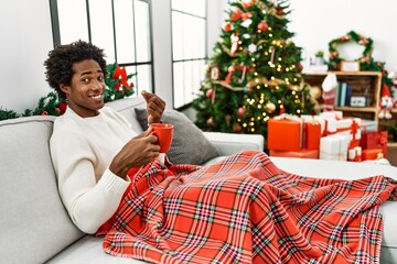 Poster - Young african american man sitting on the sofa drinking coffee by christmas tree doing money gesture with hands, asking for salary payment, millionaire business