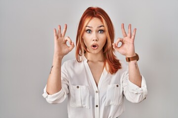 Sticker - Young caucasian woman standing over isolated background looking surprised and shocked doing ok approval symbol with fingers. crazy expression