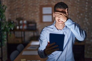 Wall Mural - Handsome hispanic man working at the office at night smiling and laughing with hand on face covering eyes for surprise. blind concept.