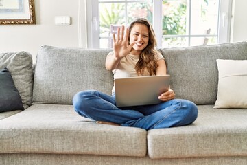 Sticker - Beautiful young brunette woman sitting on the sofa using computer laptop at home showing and pointing up with fingers number five while smiling confident and happy.