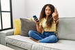 Beautiful hispanic woman sitting on the sofa at home using smartphone showing middle finger, impolite and rude fuck off expression