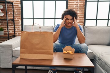 Poster - Hispanic man with curly hair eating chicken wings with hand on head, headache because stress. suffering migraine.