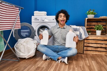 Wall Mural - Hispanic man with curly hair doing laundry holding detergent bottles celebrating crazy and amazed for success with open eyes screaming excited.