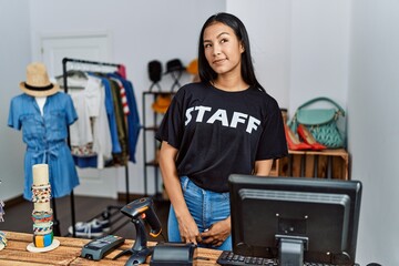 Wall Mural - Young hispanic woman working as staff at retail boutique smiling looking to the side and staring away thinking.