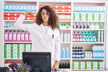 Hispanic woman with curly hair working at pharmacy drugstore strong person showing arm muscle, confident and proud of power