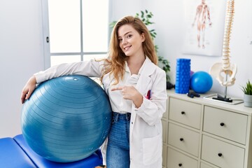 Canvas Print - Young caucasian woman working at rehabilitation clinic holding pilates ball smiling happy pointing with hand and finger