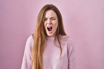 Wall Mural - Young caucasian woman standing over pink background angry and mad screaming frustrated and furious, shouting with anger. rage and aggressive concept.