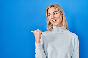 Poster - Young caucasian woman standing over blue background smiling with happy face looking and pointing to the side with thumb up.