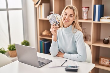 Poster - Young caucasian woman using laptop holding dollars banknotes looking positive and happy standing and smiling with a confident smile showing teeth