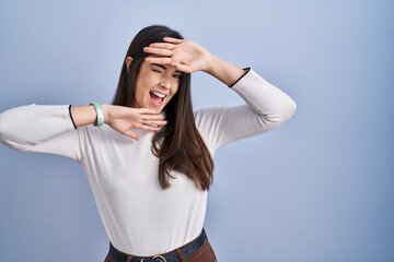 Poster - Young brunette woman standing over blue background smiling cheerful playing peek a boo with hands showing face. surprised and exited