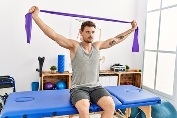 Sticker - Young hispanic man stretching arm using elastic band sitting on massage board at clinic