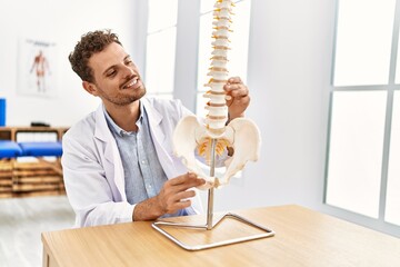 Sticker - Young hispanic man wearing physiotherapist uniform touching anatomical model of vertebral column at clinic