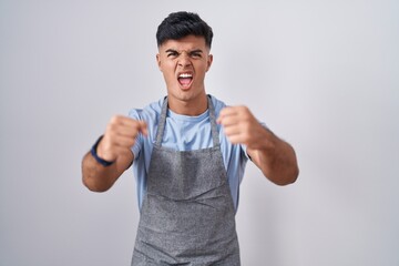 Canvas Print - Hispanic young man wearing apron over white background angry and mad raising fists frustrated and furious while shouting with anger. rage and aggressive concept.