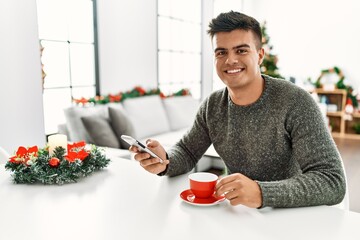 Wall Mural - Young hispanic man drinking coffee and using smartphone sitting by christmas tree at home