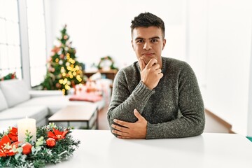 Canvas Print - Young hispanic man sitting on the table by christmas tree looking confident at the camera with smile with crossed arms and hand raised on chin. thinking positive.