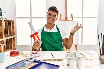 Poster - Young hispanic man at art studio holding degree smiling cheerful presenting and pointing with palm of hand looking at the camera.