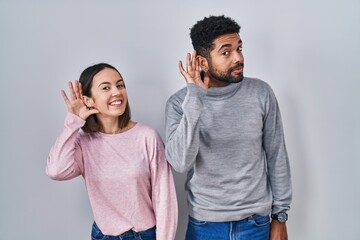 Poster - Young hispanic couple standing together smiling with hand over ear listening an hearing to rumor or gossip. deafness concept.