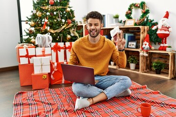 Poster - Arab young man using laptop sitting by christmas tree smiling with happy face winking at the camera doing victory sign with fingers. number two.