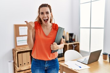 Poster - Young redhead woman at the office holding clipboard pointing thumb up to the side smiling happy with open mouth