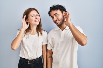 Wall Mural - Young couple wearing casual clothes standing together smiling with hand over ear listening an hearing to rumor or gossip. deafness concept.