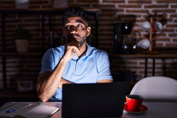 Wall Mural - Hispanic man with beard using laptop at night with hand on chin thinking about question, pensive expression. smiling with thoughtful face. doubt concept.