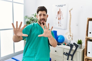 Wall Mural - Young man with beard working at pain recovery clinic afraid and terrified with fear expression stop gesture with hands, shouting in shock. panic concept.