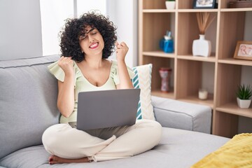 Sticker - Young brunette woman with curly hair using laptop sitting on the sofa at home very happy and excited doing winner gesture with arms raised, smiling and screaming for success. celebration concept.