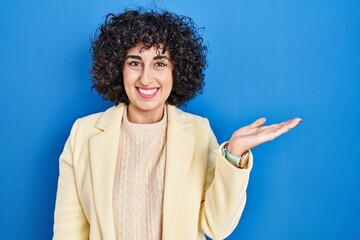 Sticker - Young brunette woman with curly hair standing over blue background smiling cheerful presenting and pointing with palm of hand looking at the camera.