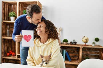 Sticker - Middle age hispanic couple kissing and holding paper with heart draw at home.