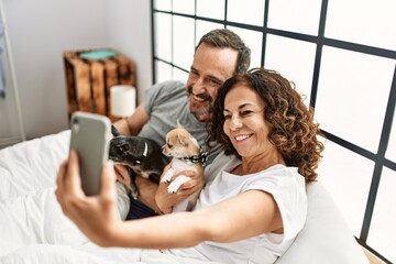 Sticker - Middle age hispanic couple smiling happy making selfie by the smartphone. Lying on the bed with dogs at home.