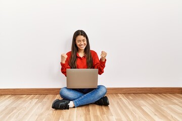 Canvas Print - Young brunette woman sitting on the floor at empty room with laptop very happy and excited doing winner gesture with arms raised, smiling and screaming for success. celebration concept.