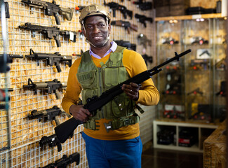 African-american man wearing armor and helmet while standing with assault rifle in airsoft shop.