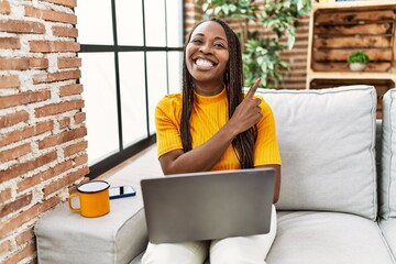 Poster - African woman sitting on the sofa using laptop at home smiling happy pointing with hand and finger to the side