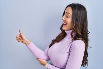 Poster - Young brunette woman standing over blue background looking proud, smiling doing thumbs up gesture to the side